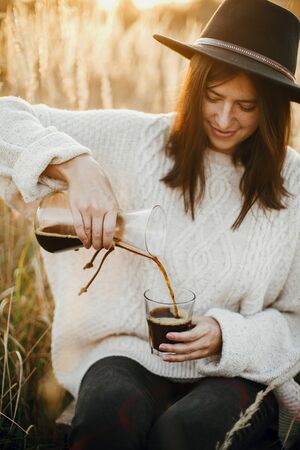 Hipster woman pouring hot coffee in glass cup on background of rural herbs in sunset. Alternative coffee brewing outdoors in sunny light. Atmospheric rustic moment. Wanderlustの写真素材