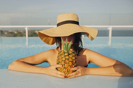 Beautiful young woman in hat drinking delicious cocktail from pineapple and relaxing in pool, summer vacation. Girl enjoying warm sunshine in pool on rooftop in luxury tropical resortの写真素材