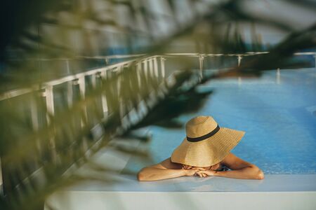 Stylish young woman in hat relaxing in pool under palm leaves and enjoying warm sunny  summer...