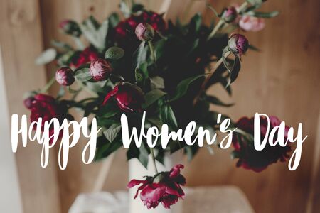 Happy women's day text on stylish peony bouquet in white vase on rustic wooden background. Red and...