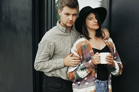 Stylish hipster couple with morning coffee relaxing on background of modern cabin and big windows in mountains. Happy young family of homeowners, enjoying morning in new home in woodsの写真素材