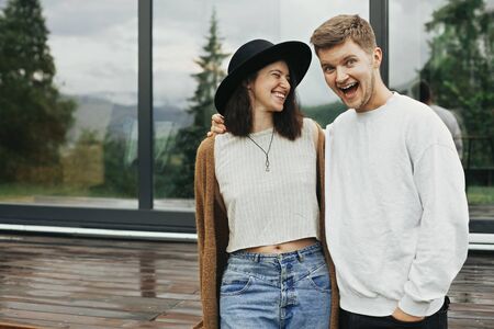 Stylish hipster couple smiling on wooden porch, relaxing in modern cabin in mountains. Happy young...