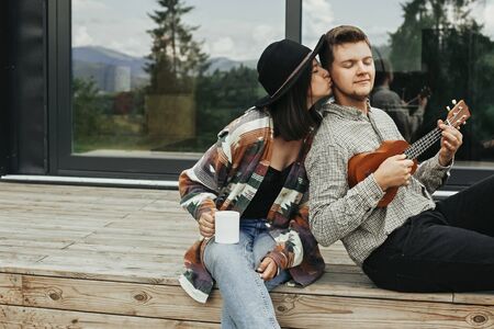 Happy hipster couple relaxing on wooden porch of modern cabin with big windows in mountains. Hipster man playing on ukulele for his stylish woman, enjoying new home in woods.の写真素材
