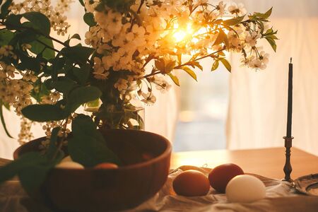 Happy Easter, atmospheric moment. Natural Easter eggs and vintage candle in sunset warm light on linen rustic fabric with cherry flowers and green leaves. Rural still life. White and brown eggsの写真素材