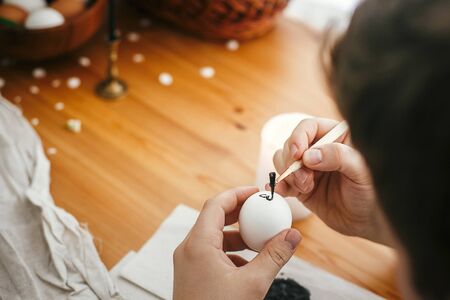 Hands painting easter egg with hot wax on background of wooden table. Modern Easter eggs with...