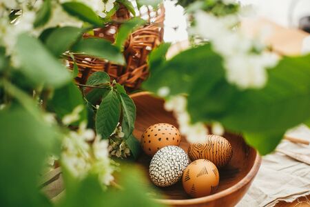 Natural Easter eggs painted with wax in wooden bowl on background of wicker basket and white spring flowers on wooden table. Happy Easter. Rural still life. Eco holiday. Zero wasteの写真素材