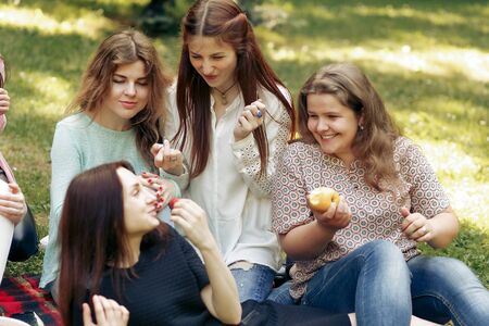 stylish happy group of women eating fruits and having fun smiling on picnic, joyful moments in summer parkの写真素材