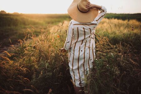 Woman in rustic dress and hat walking in wildflowers and herbs in sunset golden light in summer meadow. Atmospheric authentic moment. Stylish girl enjoying evening in countryside.の写真素材