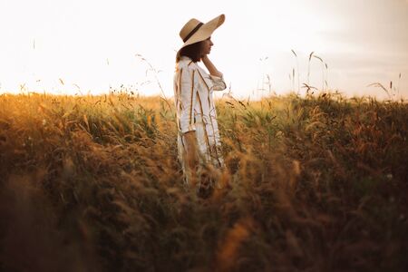 Woman in rustic dress and hat walking in wildflowers and herbs in sunset golden light in summer meadow. Stylish girl enjoying evening in countryside. Rural slow life. Copy spaceの写真素材