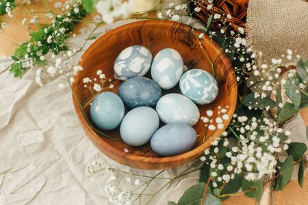 Modern Easter eggs in wooden bowl with spring flowers and eucalyptus on rustic table. Stylish pastel...