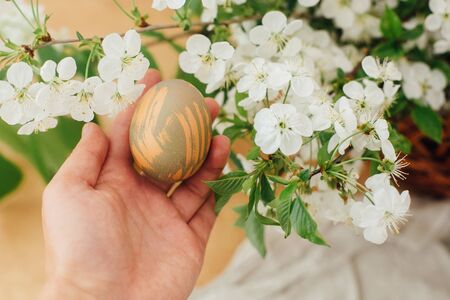 Happy Easter. Hand holding modern Easter egg on background of blooming spring flowers on rustic...