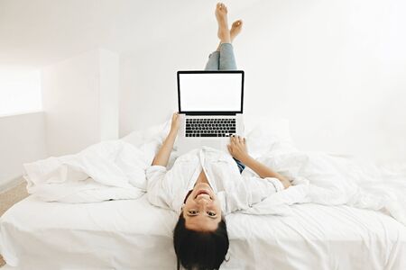 Happy stylish girl lying on white bed with laptop empty screen and smiling. Joyful young woman...