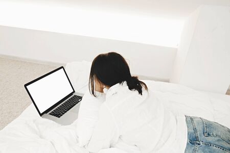 Stylish girl lying on bed with laptop with blank screen in white modern room. Young woman looking at empty display, shopping online or working online from home template. Freelanceの写真素材