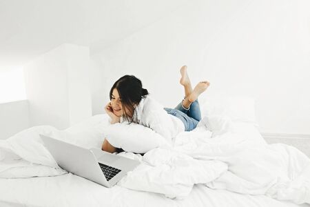 Happy stylish girl looking at laptop, lying on bed in white modern room. Young casual woman shopping online or studying online from home and smiling. Stay home. Freelanceの写真素材