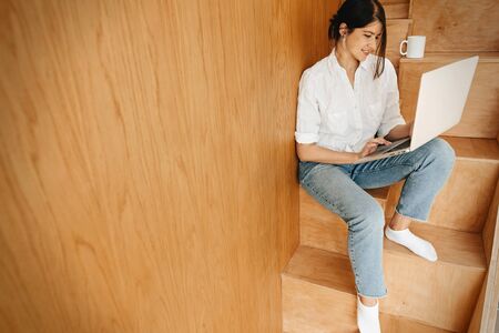 Happy stylish girl using laptop, sitting on wooden stairs with cup of coffee. Young casual woman shopping online or working online from home and smiling. Stay home. Freelanceの写真素材