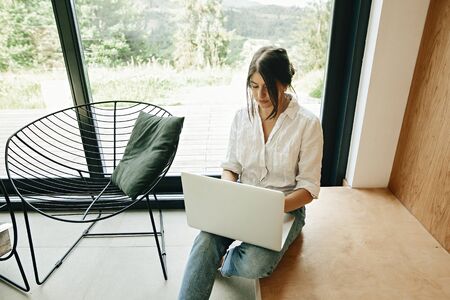 Stylish hipster girl with laptop sitting on floor at big window in modern room. Young woman using...