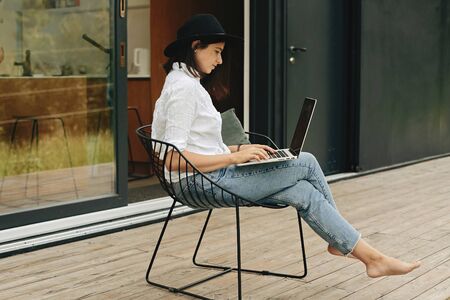 Stylish hipster girl sitting with laptop on wooden porch at big window. Young happy woman in hat...
