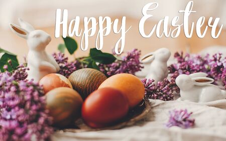 Happy Easter text. Easter greetings card. Stylish Easter eggs, white bunnies and lilac flowers on...