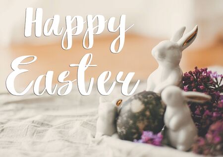 Happy Easter text. Easter greetings card. Modern Easter eggs, white bunnies and lilac flowers on...
