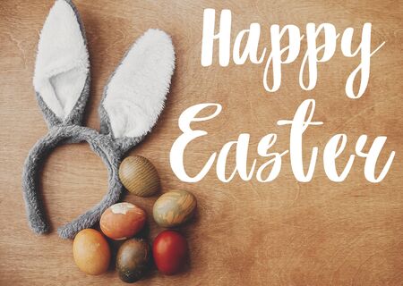 Happy Easter text. Easter greetings card. Stylish easter eggs flat lay on rustic table with bunny...