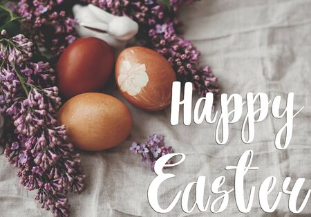 Happy Easter text. Easter greetings card. Modern colorful Easter eggs on rustic linen fabric and purple lilac flowers. Rural still life. Natural dyed easter eggs.  Holiday cardの写真素材