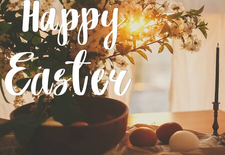 Happy Easter text. Easter greetings lettering. Natural Easter eggs in sunset warm light on linen rustic fabric with cherry blooming flowers  on  wooden table. Greeting cardの写真素材