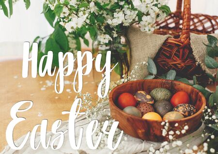 Happy Easter text. Easter greetings lettering.  Modern Easter eggs in wooden bowl with spring flowers and eucalyptus. Stylish red, green and stone eggs. Greeting cardの写真素材