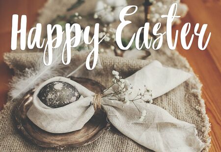 Happy Easter text. Easter greetings lettering. Stylish Easter egg in bunny ears napkin and flowers on rustic table. Modern marble egg. Holiday table decorations. Greeting cardの写真素材
