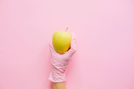 Hand in pink glove holding apple on pink background flat lay. Order groceries and get them delivered safe during quarantine. Stay home Stay safe. Prevention of virus epidemic. Clean foodの写真素材