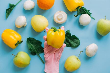 Hand in pink glove picking bell pepper on blue background with vegetables flat lay. Safe shopping in quarantine. Order food online with delivery and stay home. Prevention of virus epidemic.の写真素材