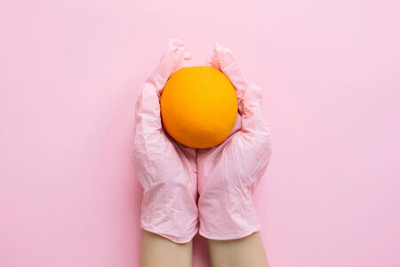 Hands in pink glove holding orange on pink background flat lay. Order groceries and get them delivered safe during quarantine. Stay home Stay safe. Prevention of virus epidemic. Clean foodの写真素材