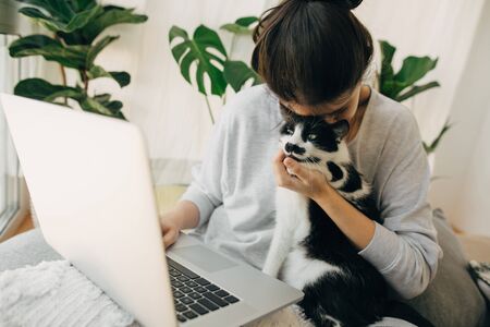Casual girl hugging her cat and working on laptop, sitting together in modern room with pillows and plants. Cute moments of friendship and happiness. Home office. Cat love. Positivityの写真素材
