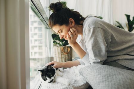 Hipster girl playing with cute cat, sitting together at home during coronavirus quarantine. Stay home stay safe. Isolation at home to prevent virus epidemic. Young woman with cat in modern roomの写真素材