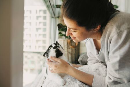 Young woman playing with cute cat, sitting together at home during coronavirus quarantine. Stay home stay safe. Isolation at home to prevent virus epidemic. Hipster girl with cat in modern roomの写真素材