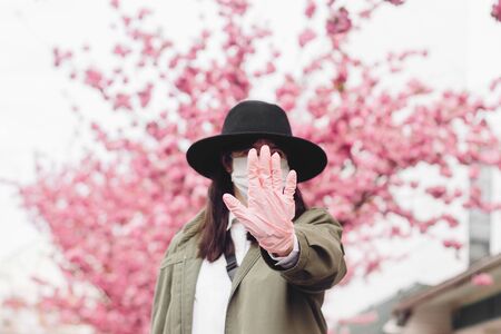 Social distancing, stay away. Woman's hand in medical glove showing stop during virus outbreak. Young girl wearing medical mask on background of blooming pink trees. Stop coronavirusの写真素材