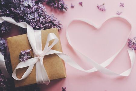 Lilac flowers, gift box and heart ribbon on pink paper, stylish flat lay. Floral greeting card. Happy mothers day concept. Purple lilac flowers bouquet with craft present boxの写真素材