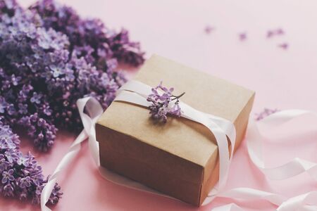 Lilac flowers and gift box on pink background. Stylish floral greeting card. Purple lilac flowers bouquet, craft present box with ribbon on pink paper. Happy mothers dayの写真素材