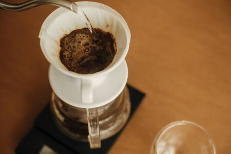 Alternative coffee brewing v60 closeup. Barista making filter coffee on brown background. Pouring hot water from steel kettle in filter with ground coffee in pour over on scale.の写真素材