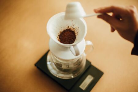 Hands pouring grounded coffee in filter pour over on glass kettle on scale. Preparing for alternative coffee brewing v60. Person holding spoon with grind coffee on brown backgroundの写真素材