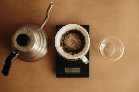 Drip coffee flat lay. Steel kettle, brewing coffee in filter pour over on scale and glass cup on brown background. Brewing aromatic fresh  alternative coffee,  v60の写真素材
