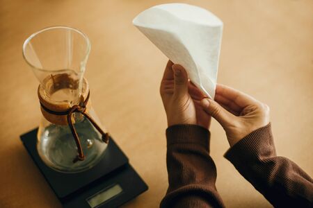 Hands folding paper filter for pour over and glass kettle on scale on brown