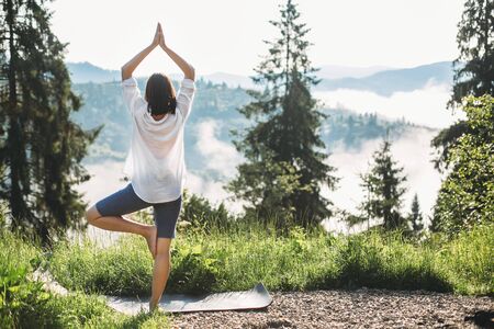 Young woman practicing yoga among grass on background of morning mountains in sunny light. Meditation. Calm tranquil moment, connection with nature. Copy spaceの写真素材