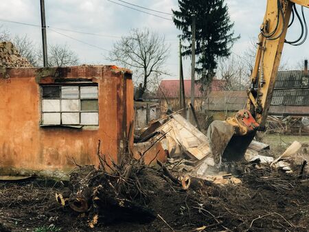 Excavator destroying brick house on land in countryside. Bulldozer clearing land from old bricks and concrete from walls with dirt and trash. Backhoe machinery ruining houseの写真素材