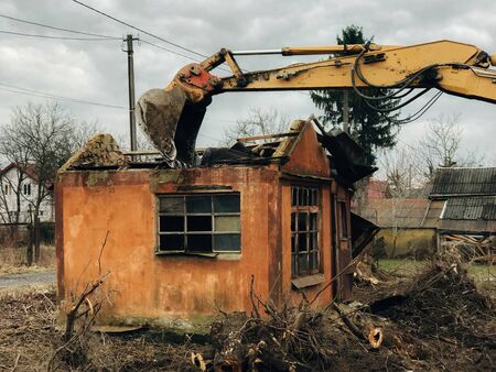 Excavator destroying brick house on land in countryside. Bulldozer clearing land from old bricks and concrete from walls with dirt and trash. Backhoe machinery ruining houseの写真素材