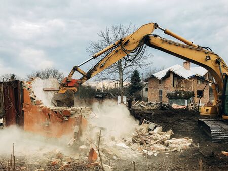 House crushing and collapse. Excavator destroying brick house on land in countryside. Bulldozer clearing land from old bricks and concrete from walls with dirt and trash. Ruining houseの写真素材