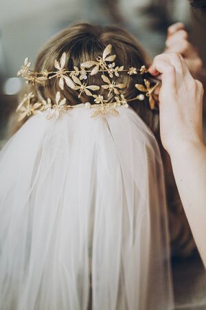 Hair stylish putting on stylish bride golden tiara with butterflies, morning preparations for wedding day. Bride in hair salon styling her hair with modern authentic wreath and veilの写真素材