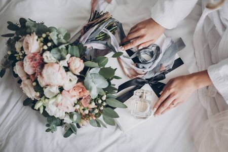 Modern wedding bouquet, perfume bottle, and gift box on white bed. Bride holding white box for morning boudoir before wedding ceremony. Wedding arrangements or bridal showerの写真素材