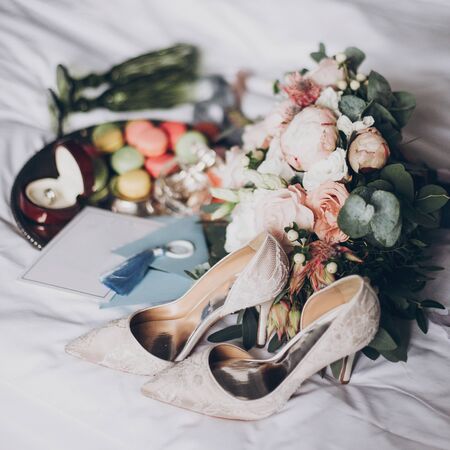Wedding preparations or bridal shower. Modern wedding bouquet, wedding rings, stylish invitation, perfume bottle, shoes, delicious macarons and green glasses for champagne on white bed.の写真素材