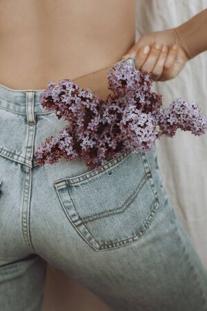 Blooming lilac flowers in back pocket. Woman posing with lilac flowers in denim jeans pocket on rustic background.  Creative image. Sensual atmospheric mood. Hello springの写真素材