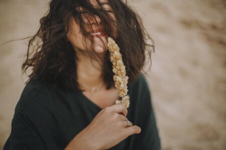 Carefree hipster girl with windy hair sitting and smiling on sandy beach, holding herb. Stylish...